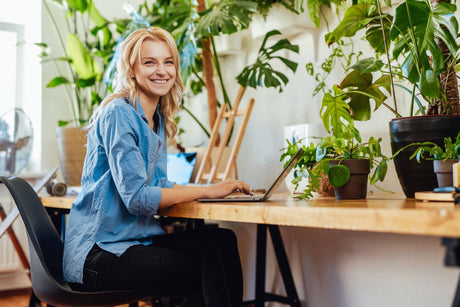 Woman working on laptop at wooden desk surrounded by lush green plants in modern home office