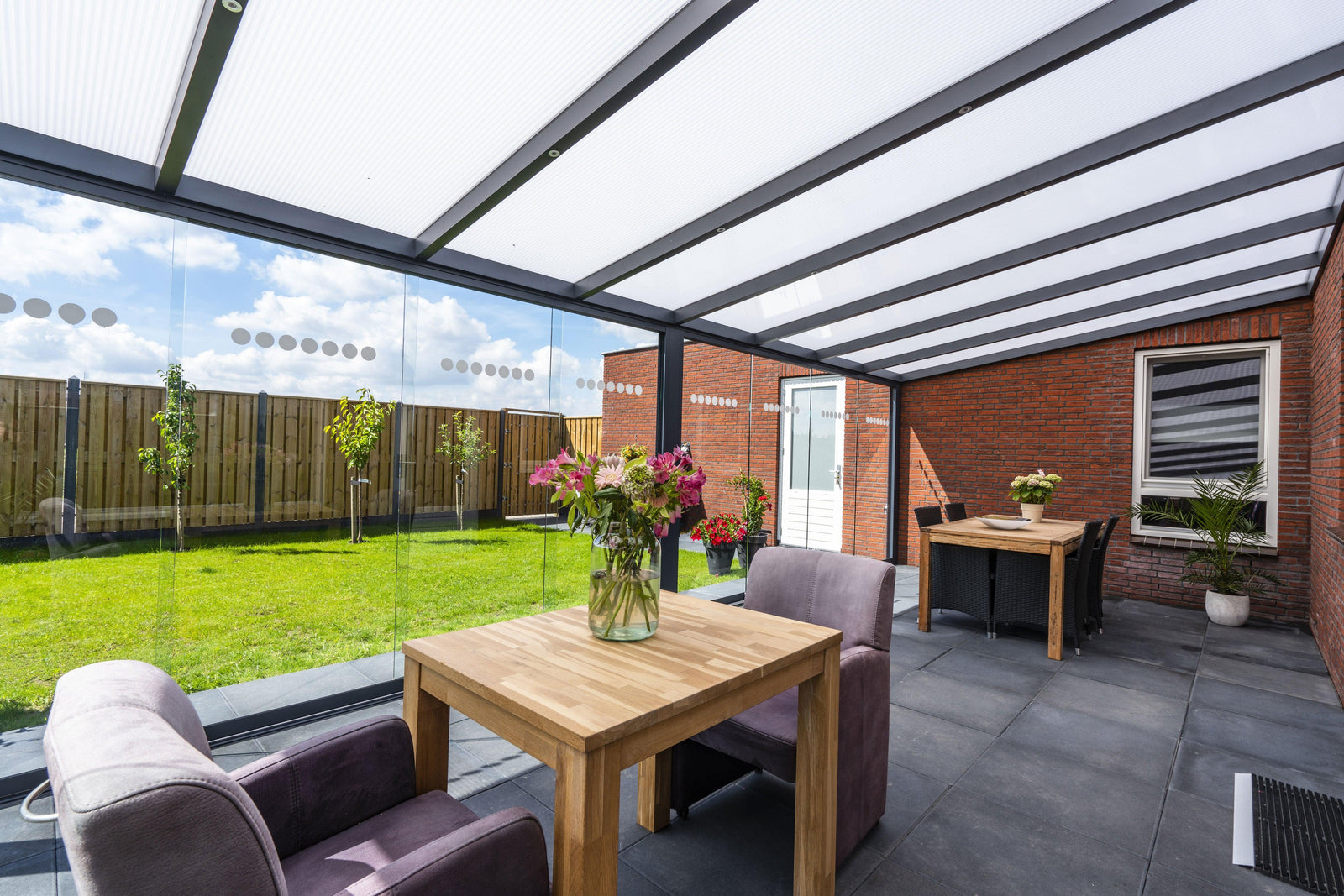 Modern glass veranda with dining area, overlooking green garden, perfect for outdoor living spaces.