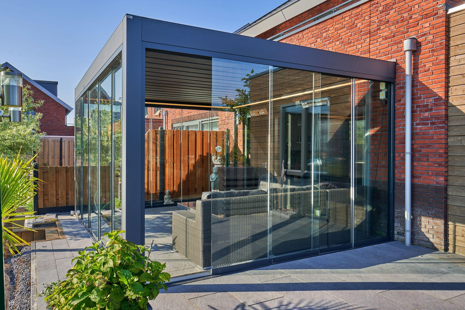 Modern glass garden room with sleek black frame, wicker sofa, and brick house backdrop for outdoor living