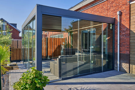 Modern glass garden room with sleek black frame, wicker sofa, and brick house backdrop for outdoor living