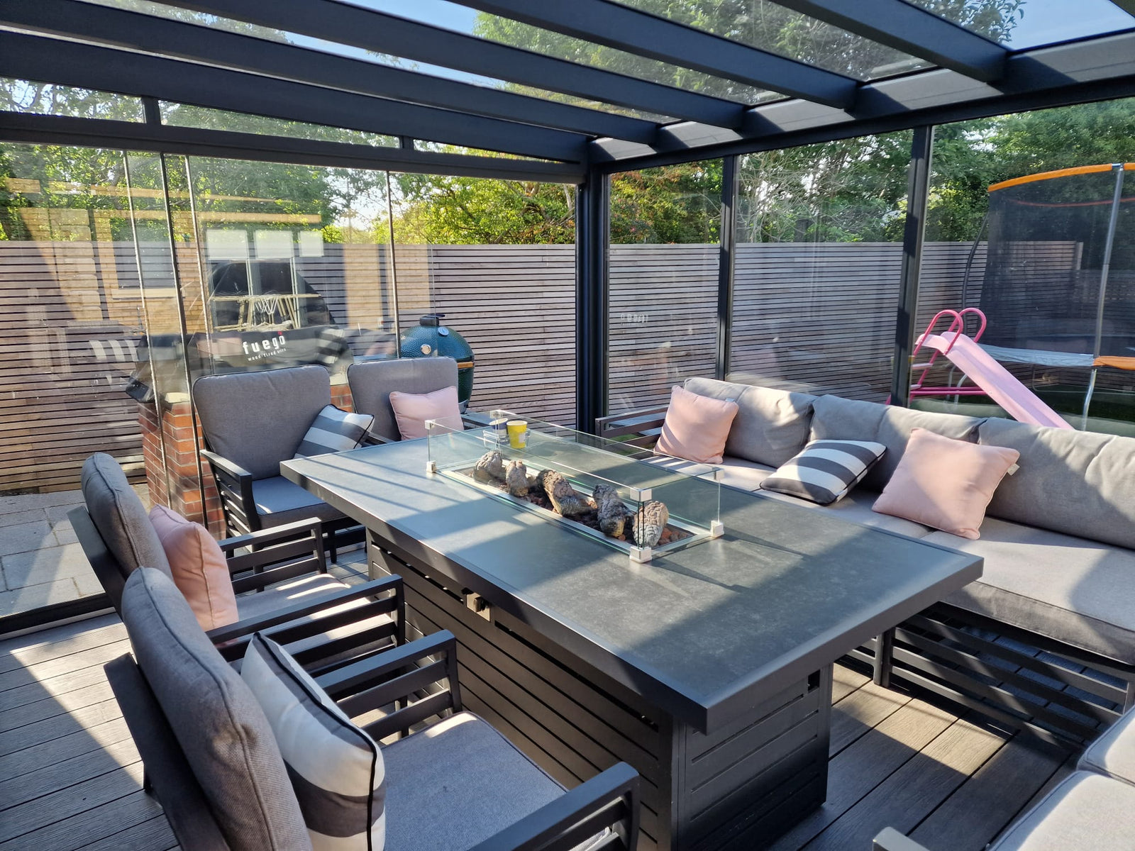Modern outdoor dining setup with fire pit table, gray seating, and glass-roofed pergola for garden living.