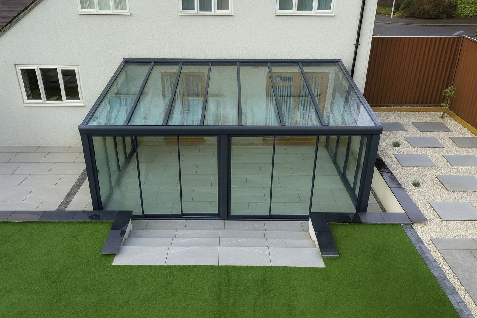 Modern glass garden room with black frame, surrounded by artificial turf and stone paving.