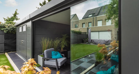 Modern garden room with gray armchair, teal pillow, and glass walls overlooking a lush green lawn.