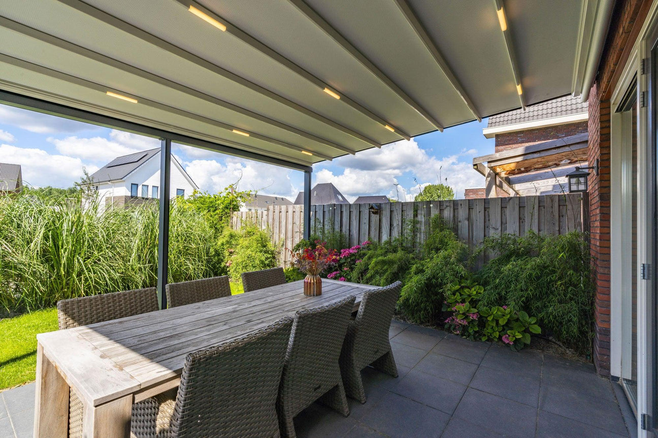 Modern outdoor dining area under a sleek pergola with integrated lighting, surrounded by lush greenery and a wooden fence.