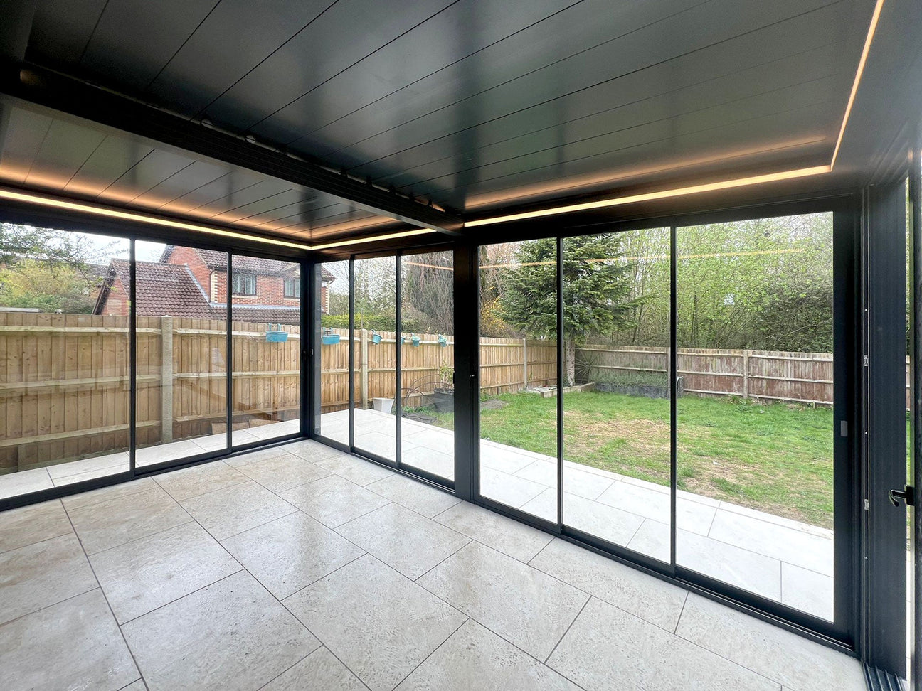 Modern glass garden room with black frame, tiled floor, and LED lighting, overlooking a fenced backyard.