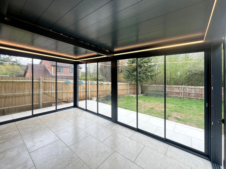 Modern glass garden room with black frame, tiled floor, and LED lighting, overlooking a fenced backyard.